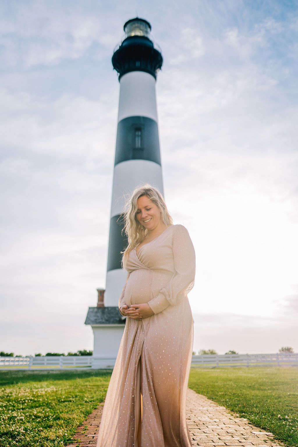 pregnant woman has the sun rising behind her as she stands in front of a light house on the beach while holding her bump