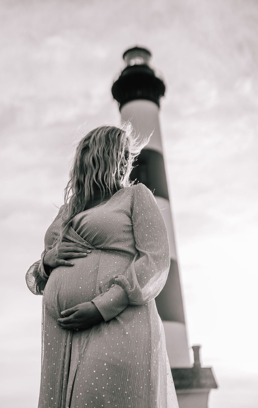 black and white photo of a pregnant woman standing in front of a lighthouse holding her bump