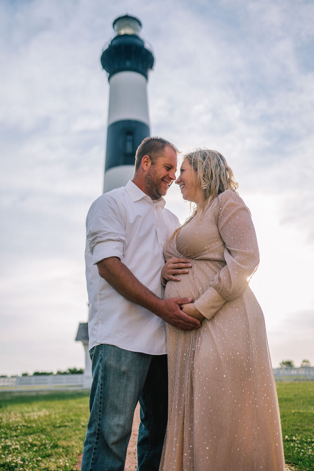 pregnant woman and her husband standing in front of a lighthouse on the beach as the hold hands for the maternity session