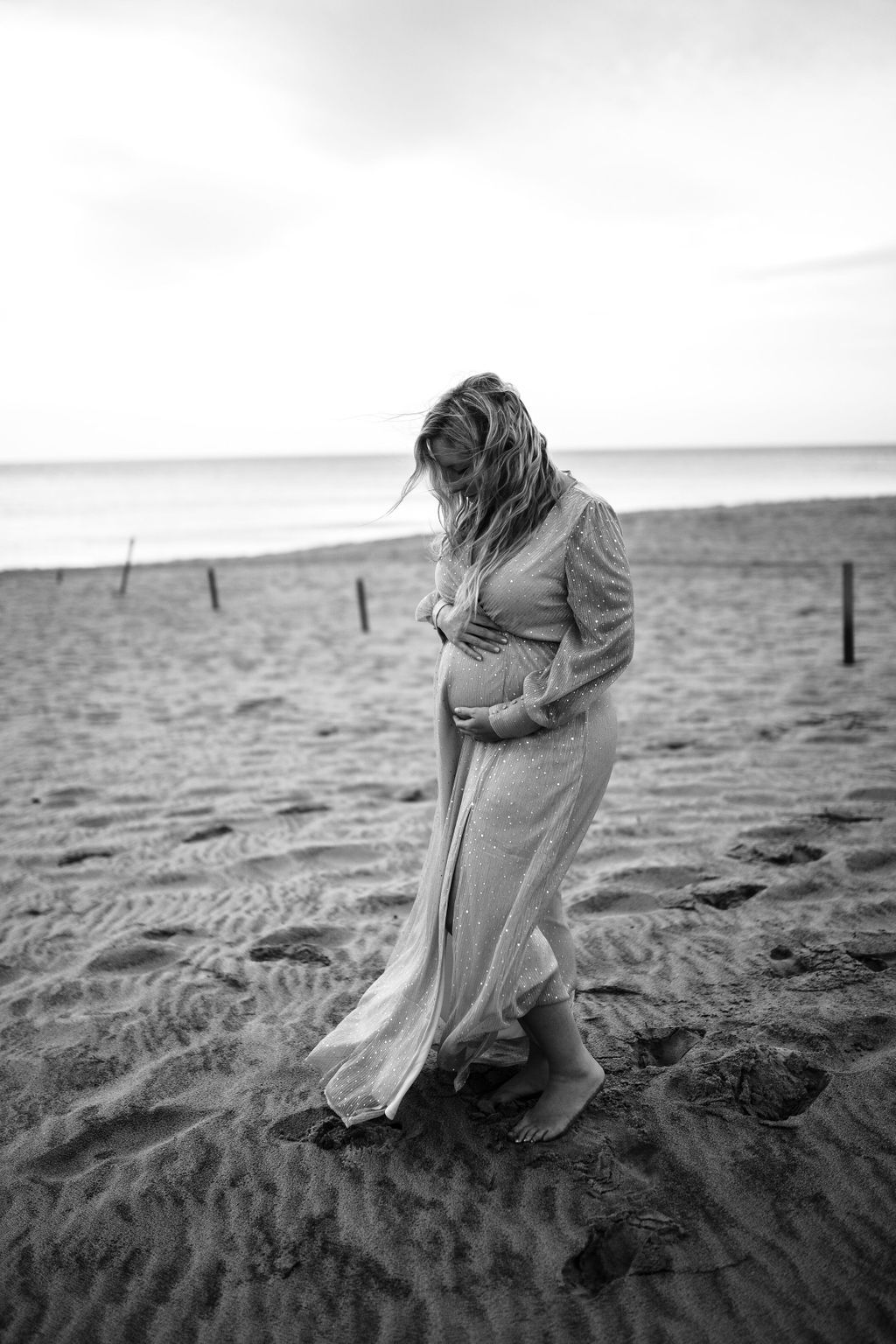 black and white photo of a woman standing on the beach at the Outerbanks holding her pregnancy bump as the wind blows her hair and dress
