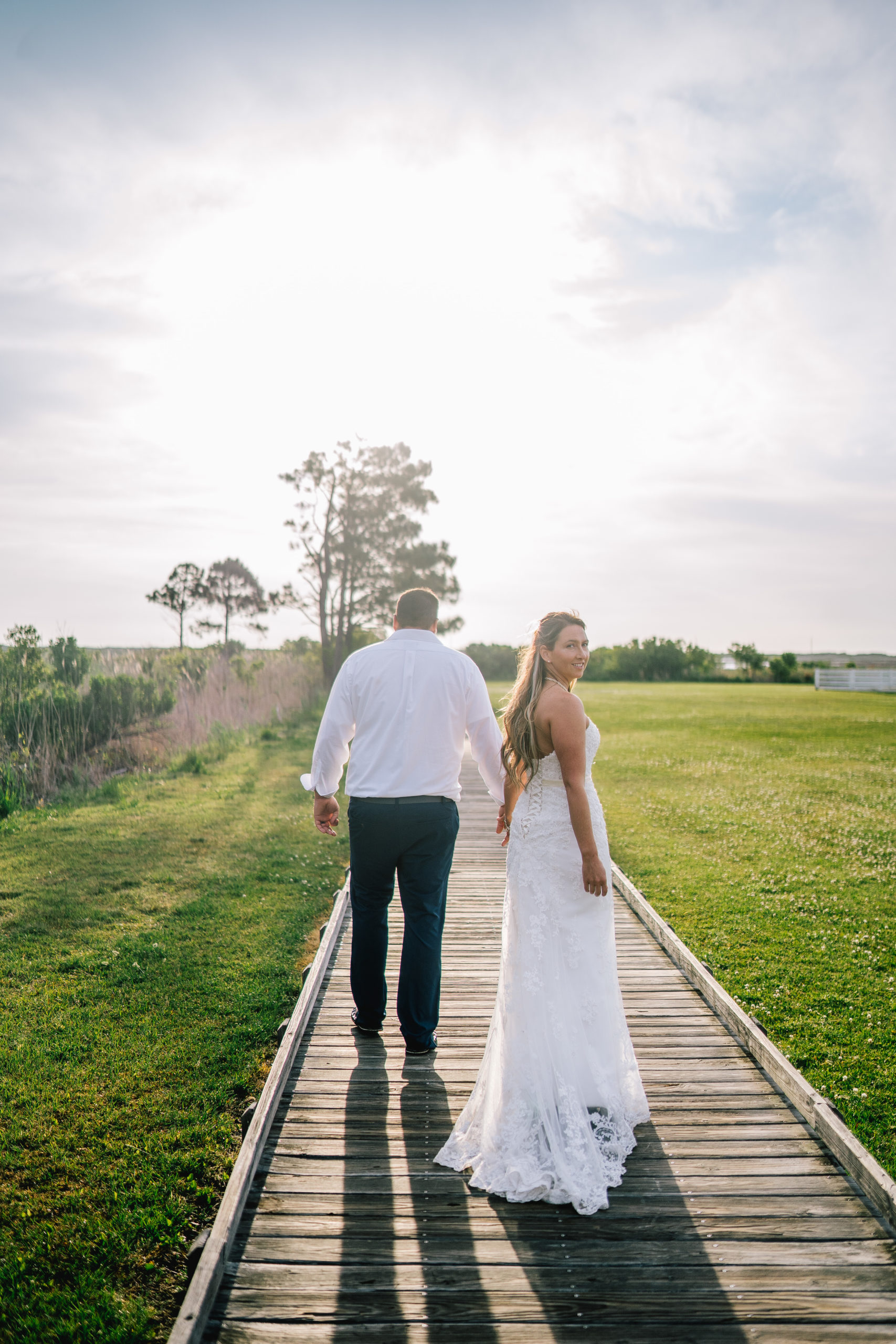 East coast couple walking on a boardwalk at sunrise holding hands as the bride who is wearing a lace dress looks behind her shoulder to the camera.