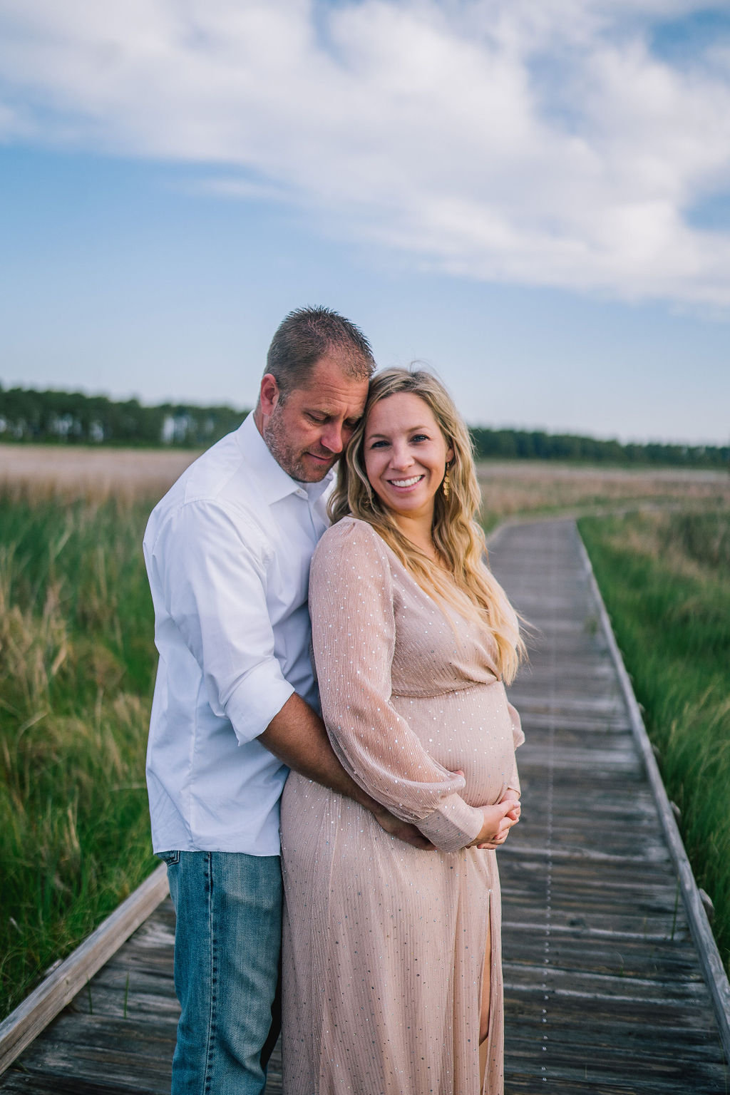 man and woman standing on a dock together, man is holding the woman's pregnant stomach and looking down at her as she smiles at the camera
