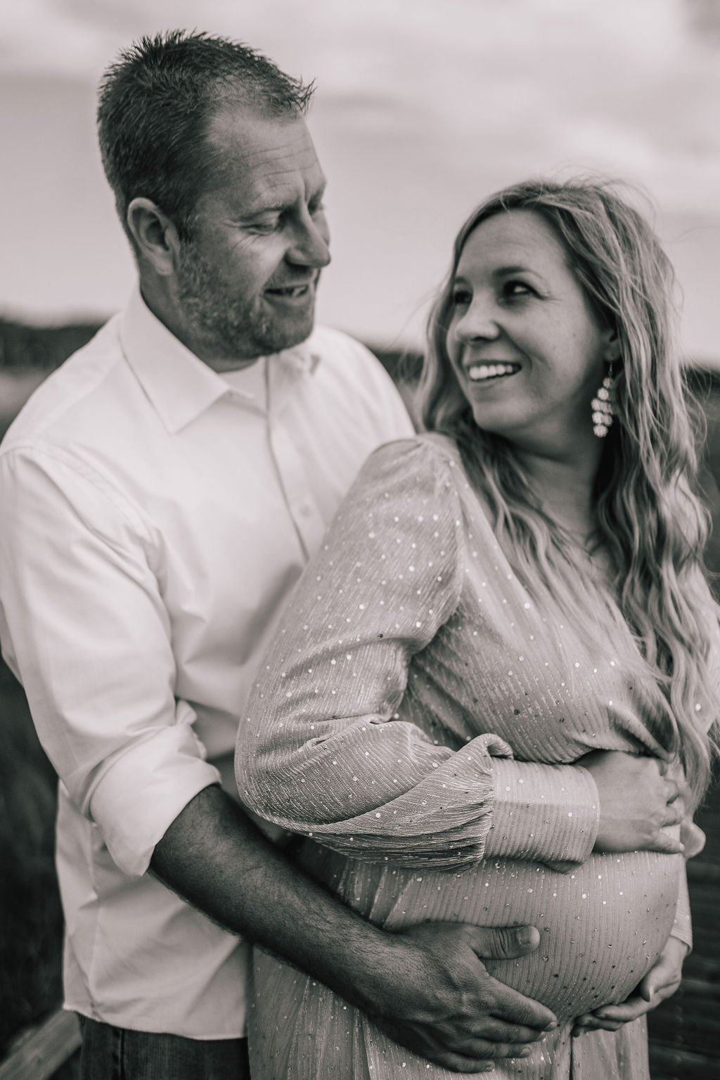 black and white photo of husband and wife embracing each other for thrit maternity session. man is holding the mothers baby bump as she looks back at him smiling.