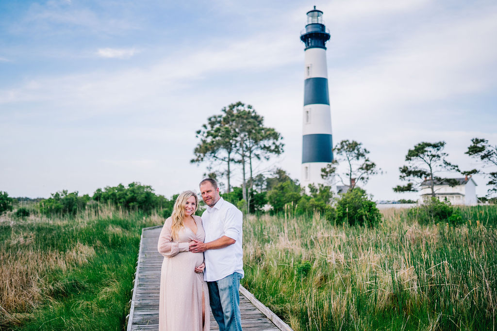 man and woman stand in front of a light house on a pier while holding the woman tummy because she is very pregnant.