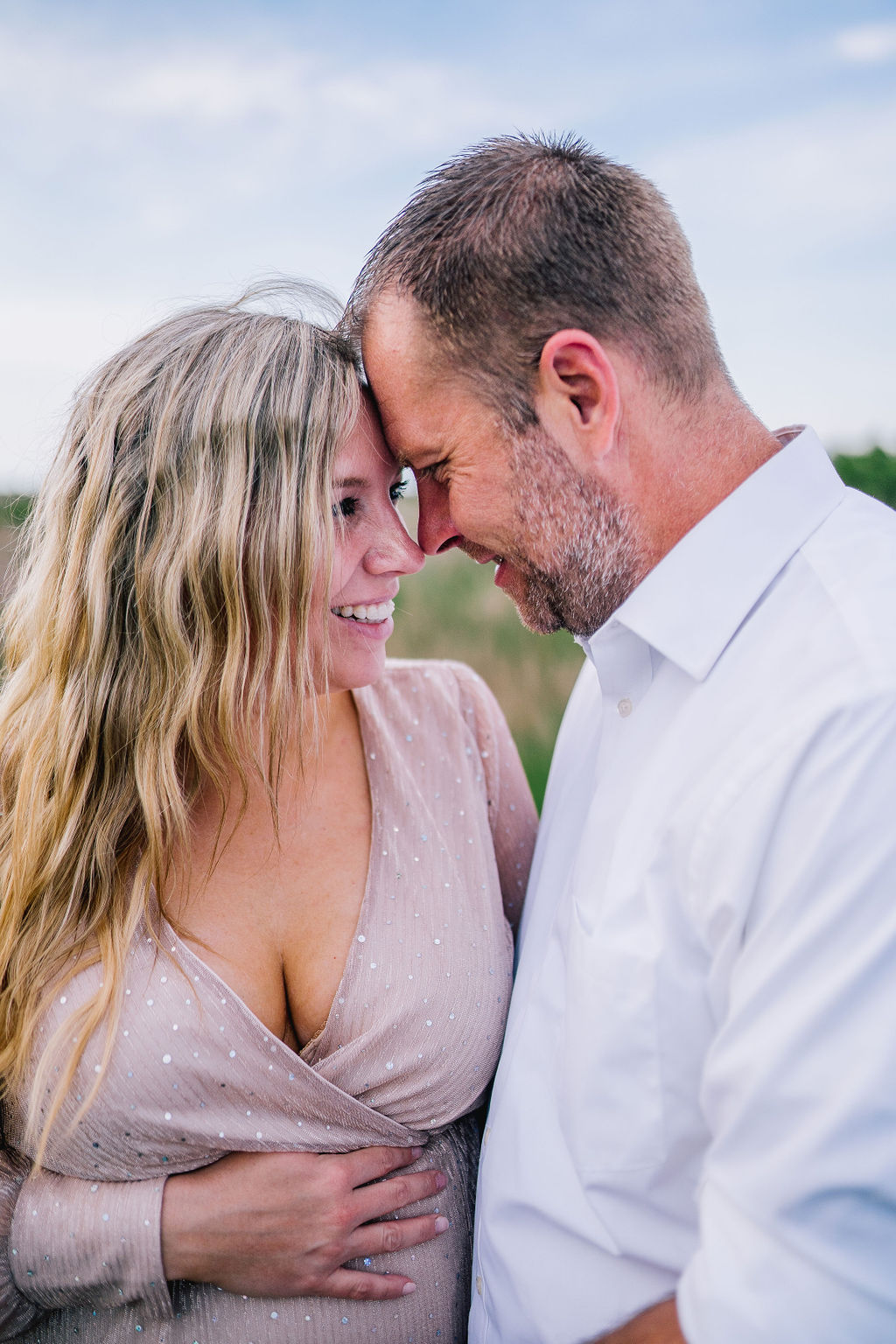 Outerbanks maternity session. Husband and wife are forehead to forehead and the woman holds her pregnancy bump in her pink formal gown.
