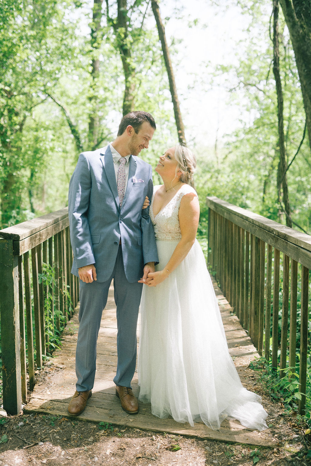 bride and groom standing on a bridge in the Ijams holding hands and gazing at each other as the light falls down on them between the leaves of the towering trees.