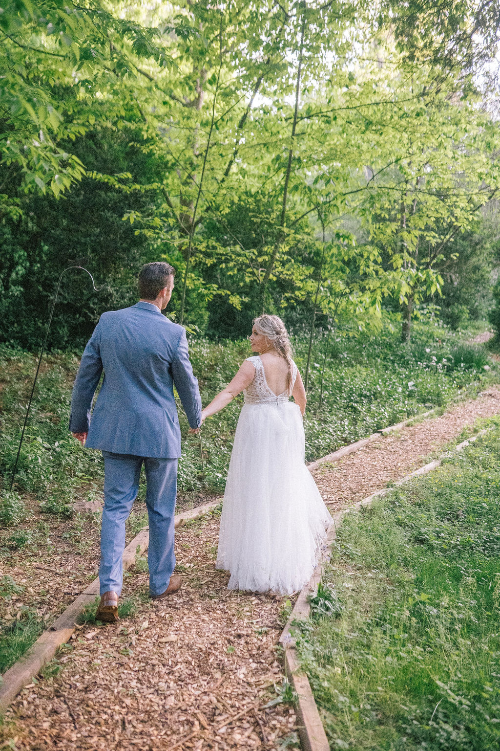 bride and groom walking on a trail in the woods of the Ijams on their wedding day, bride is wearina Vowed wedding gown and look back at her groom who is wearing a blue suit.