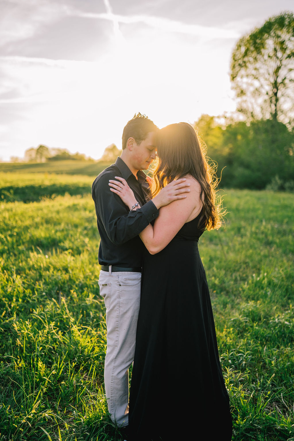 sun setting behind man and woman as they embrace each other in a grassy field with a tree line behind them in Meads Quarry Tennessee for their engagement session