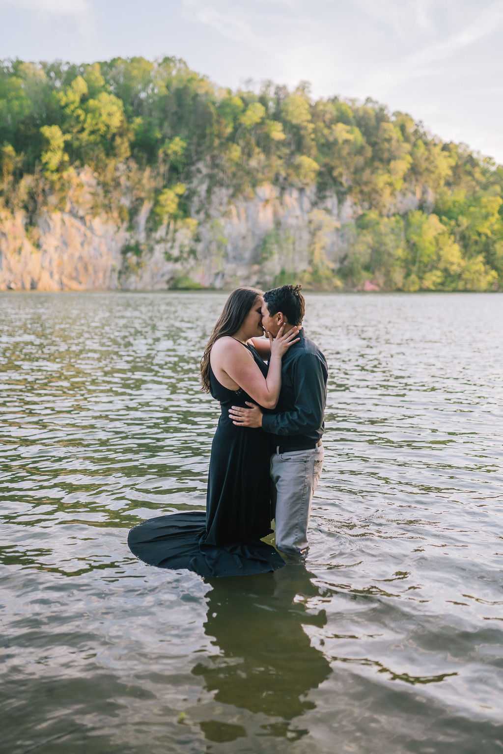 man and woman standing in the water at Meads Quarry while wearing black for their engagement session with rocks and trees behind them as they kiss