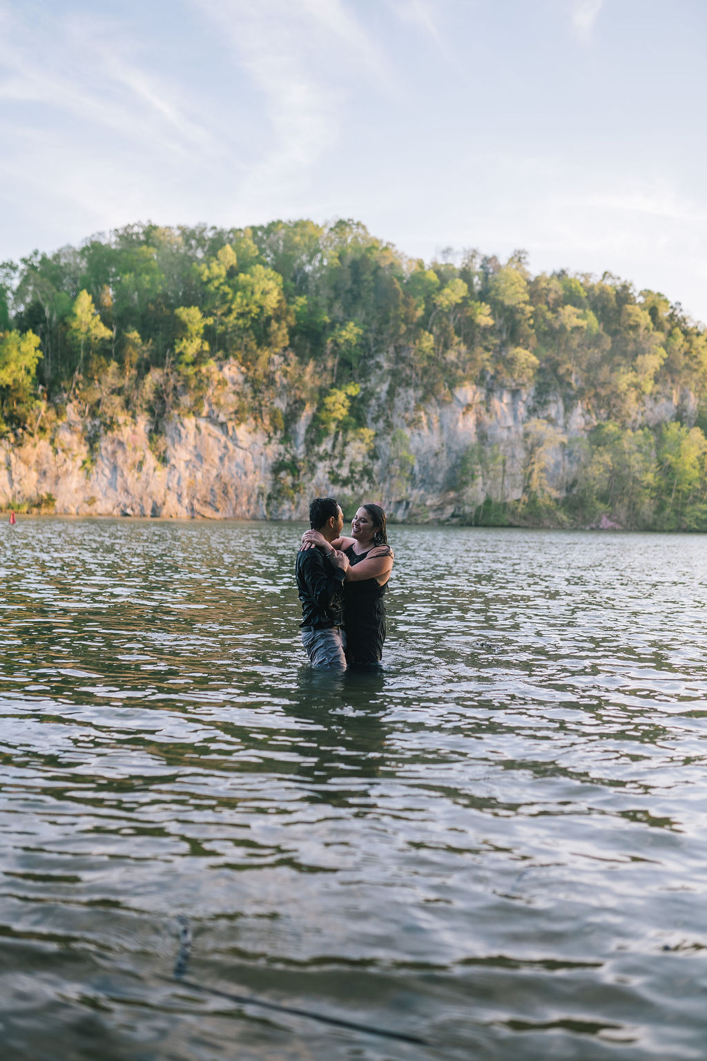 engagement session in Meads Quarry with man and woman in the water holding each other and laughing with a cliff and trees behind them
