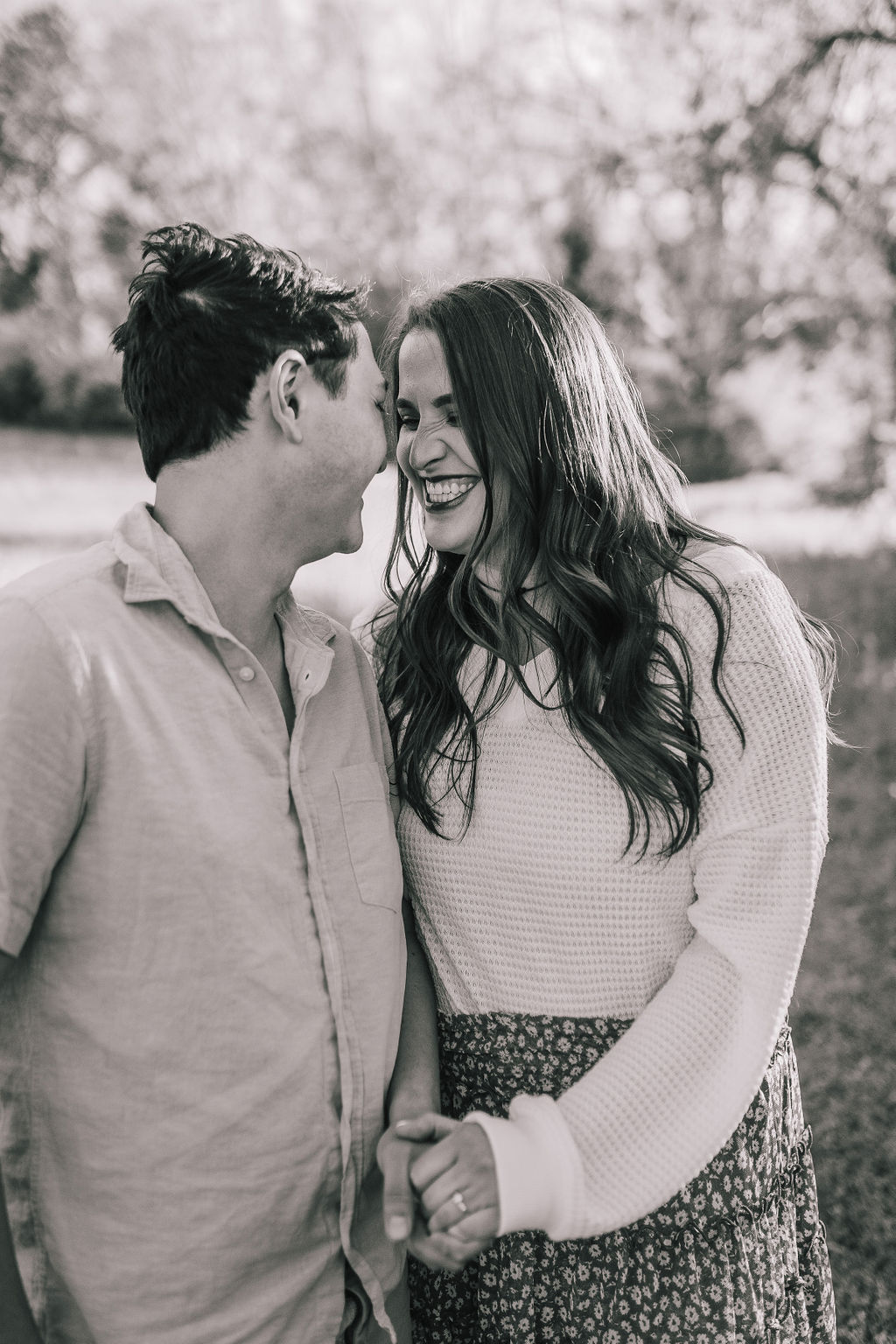 black and white engagement photo of man and woman holding hands as the man whispers into the womans ear and she laughs during their Tennessee engagement photos