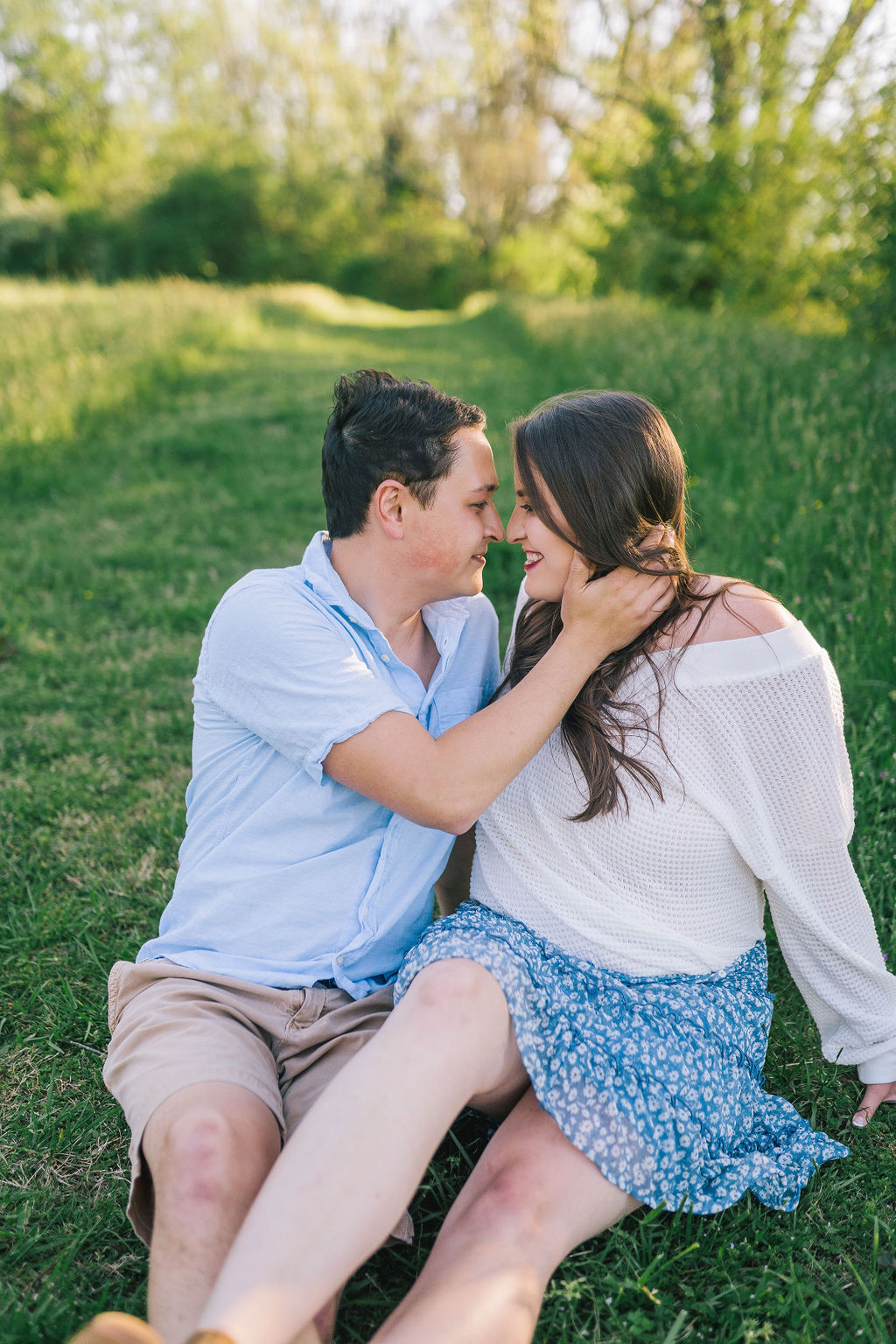 Meads Quarry engagement session with man and woman wearing blue and sitting on green grass next to a tree line, man is holding womans hair back while holding her neck as they smiles at each other