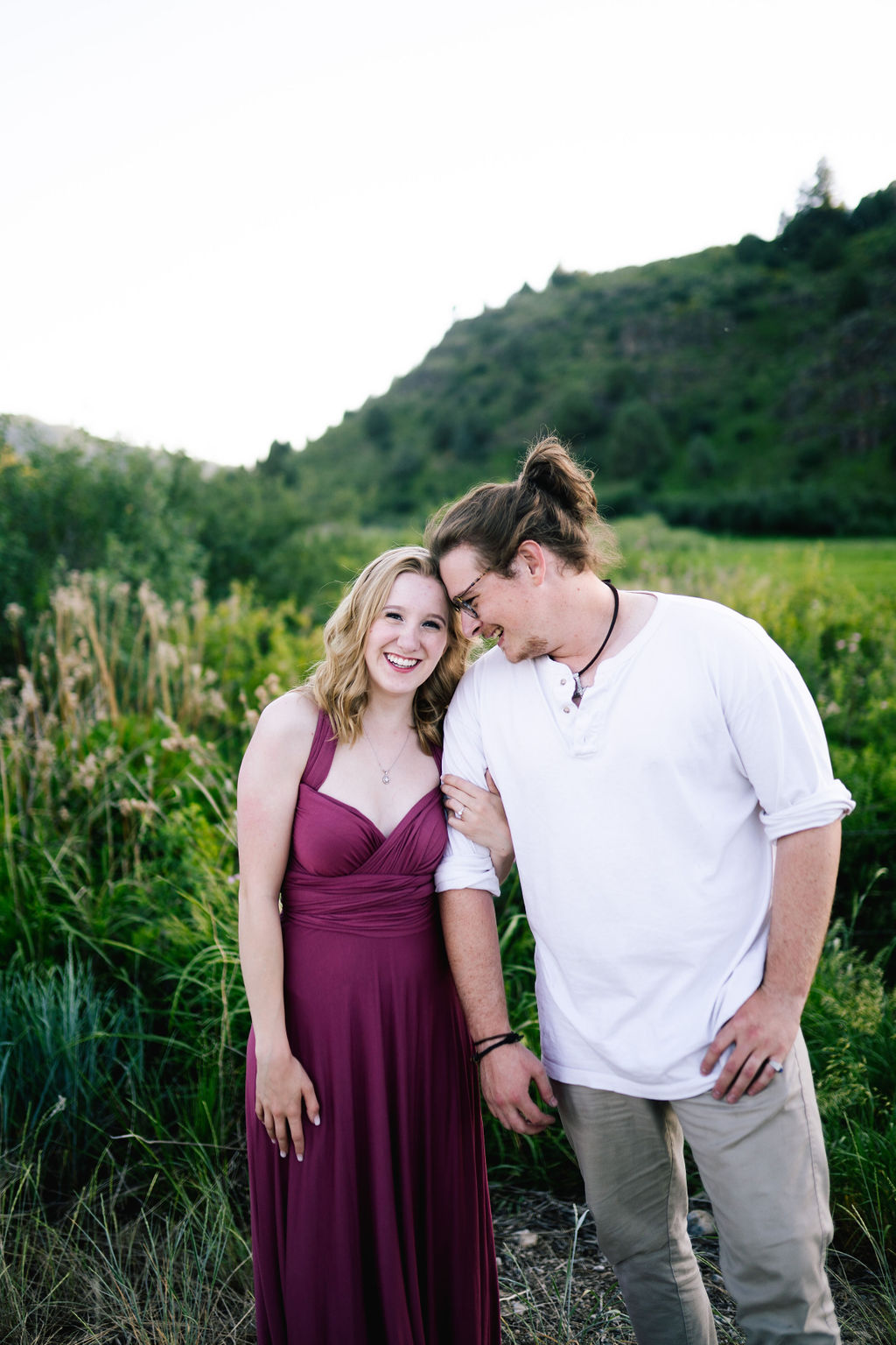 engaged couple stand together in a stunning green field and smile at the camera for their engagement photos with woma nin a purple dress and man in a white shirt looking romantically at the woman