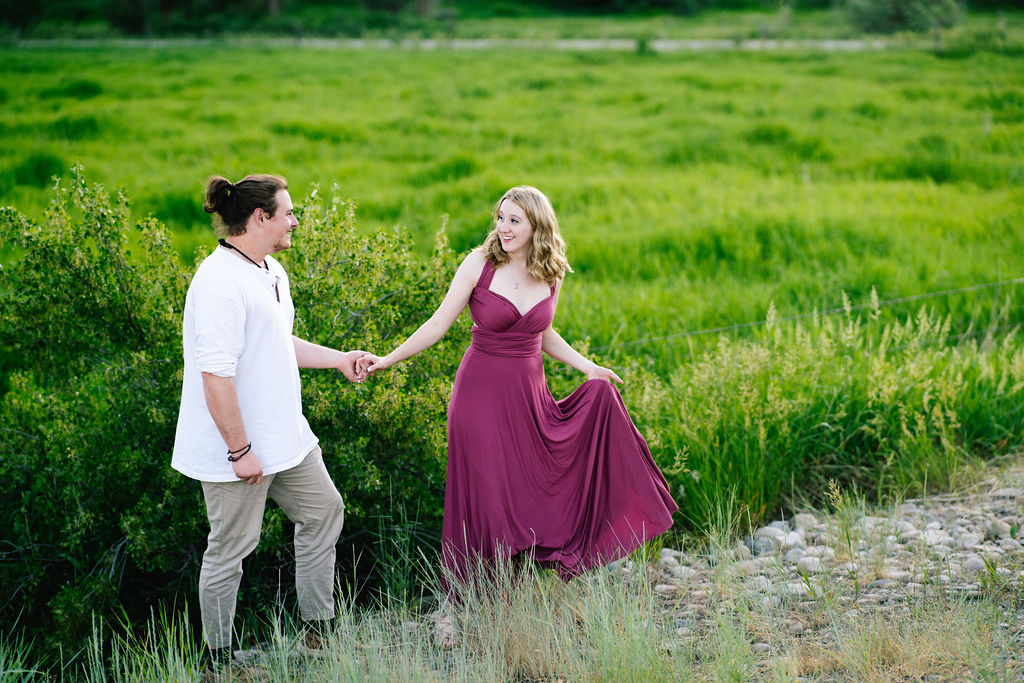woman in purple dress holds hands with her fiance and guides him through a green field in the Smokies
