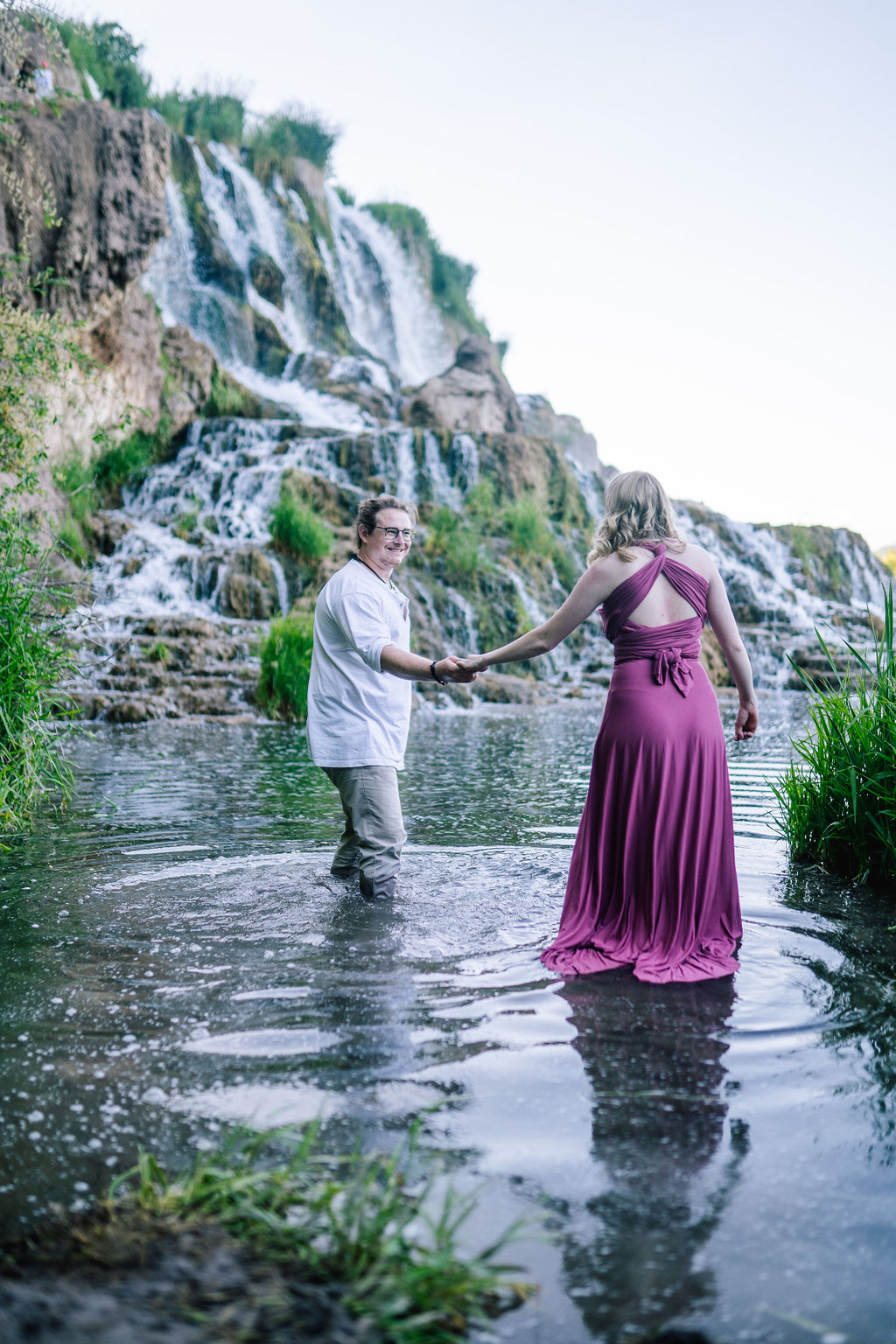 outdoor engagement photos with woman in purple dress walking into a pond with her fiance while holding hands