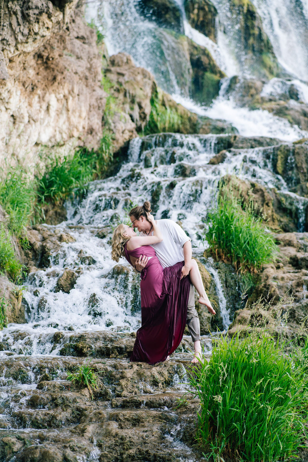 engaged couple stand on the rocks of a waterfalls together and kiss as the man dips the woman in a purple dress back and pulls her leg up