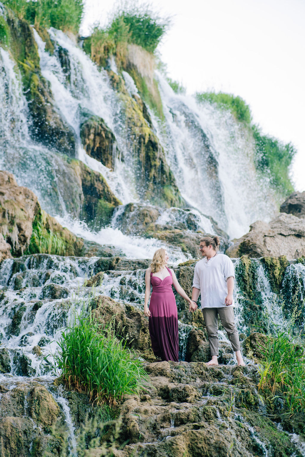 holding hands on the rocks of a waterfall the engaged couple look at each other romantically with a roaring waterfall just behind them for their waterfall engagement session