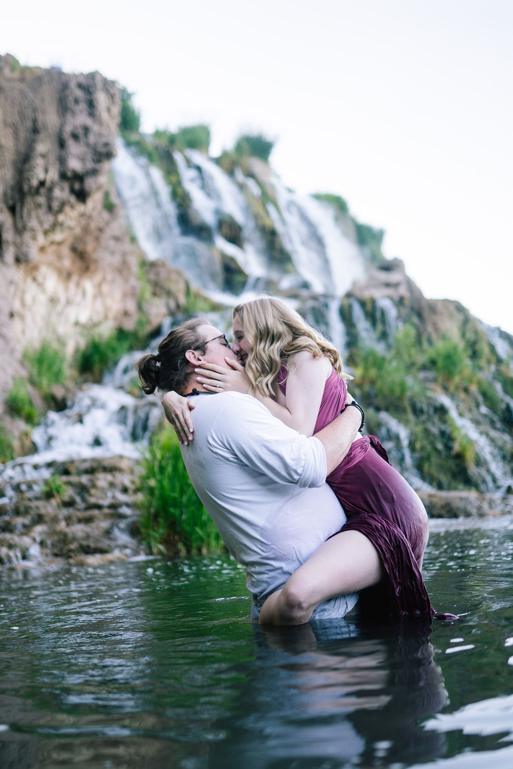 man picks up woman and woman hold back of mans neck as they kiss in a lake next to a romantic waterfall of an outdoor engagement session