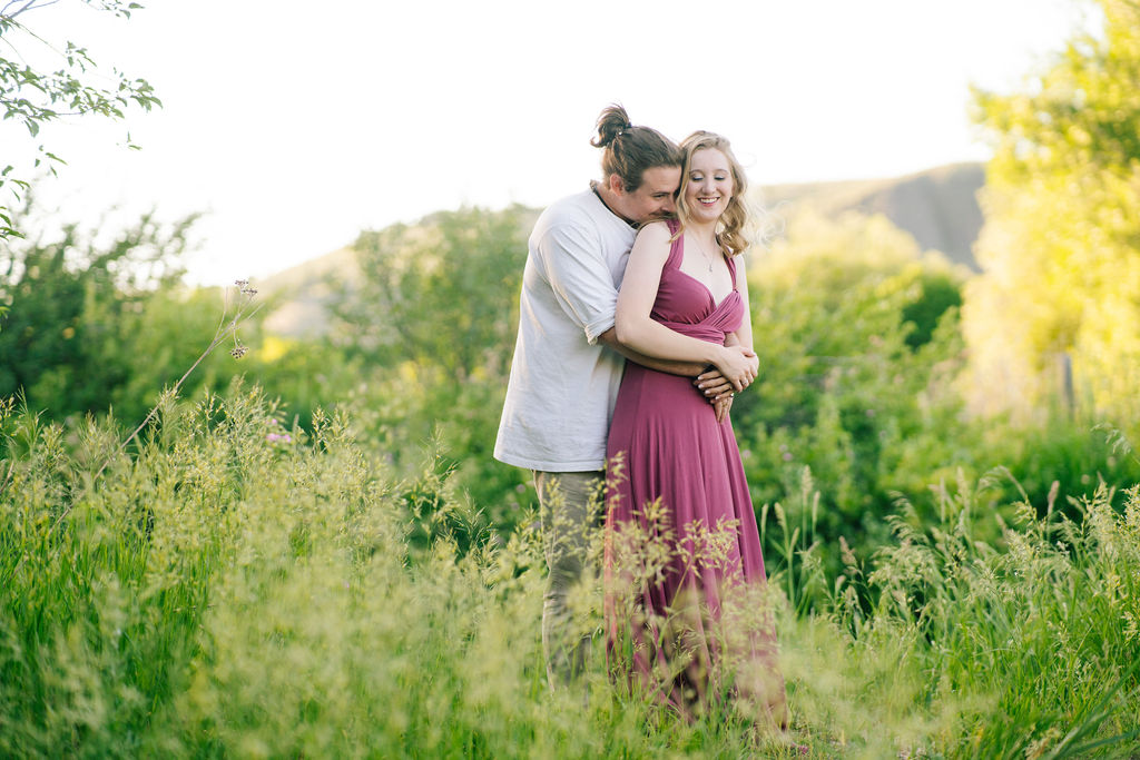 man holding his fiance from behind around her waist an kisses her shoulder as she giggles and the sun set behind them through the trees during their Knoxville Tennessee engagement session
