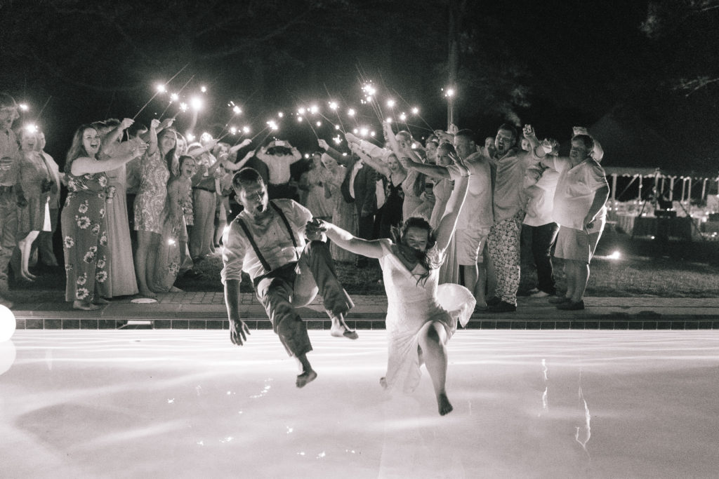 black and white image of bride and groom jumping into a pool at the end of their wedding day with their guests behind them waving sparklers in the air and cheering
