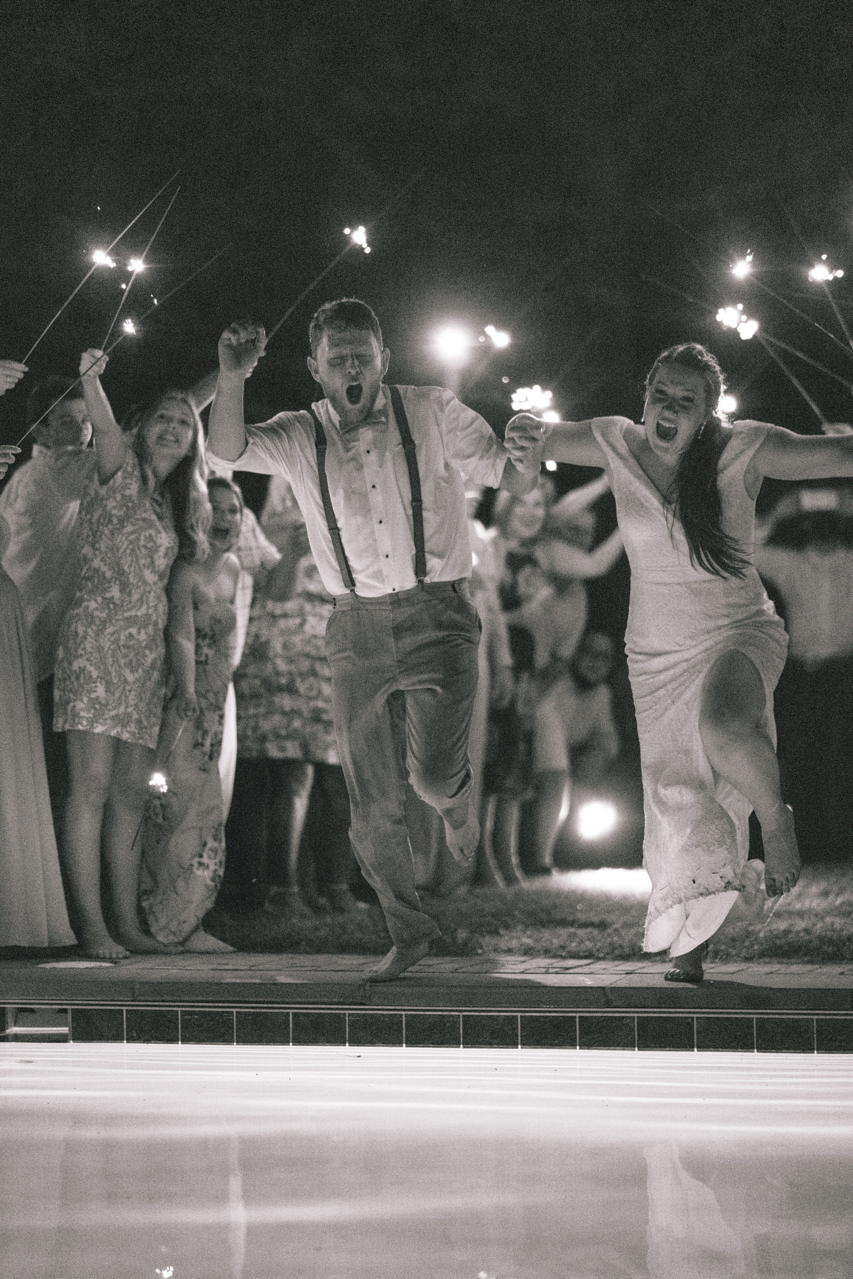 black and white image of bride and groom running through a tunnel of sparkler and jumping into a pool on their wedding day for their send off