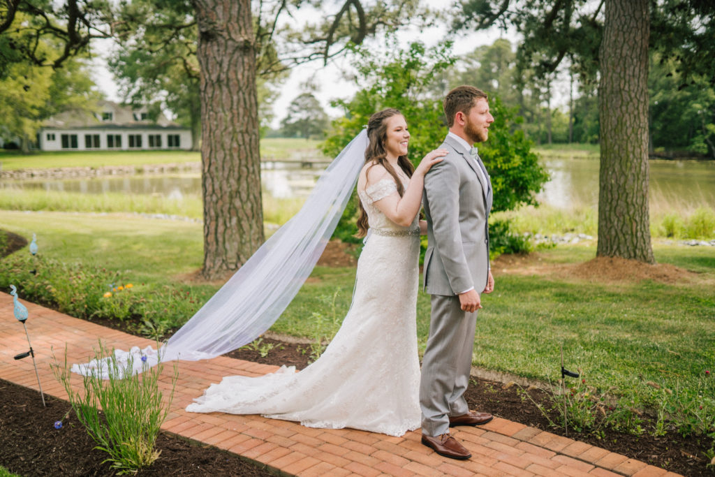 Maryland wedding with bride behind her groom tapping on her shoulder for a first look as they stand next to a river and tall trees tower over them