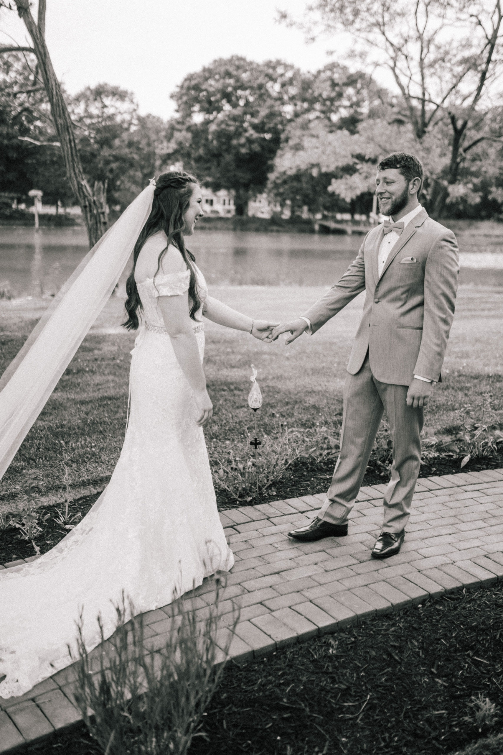 black and white picture of bride holding her grooms hand as he turns around to look at his bride for the first time with a lake behind the couple as they smile with tears in their eyes
