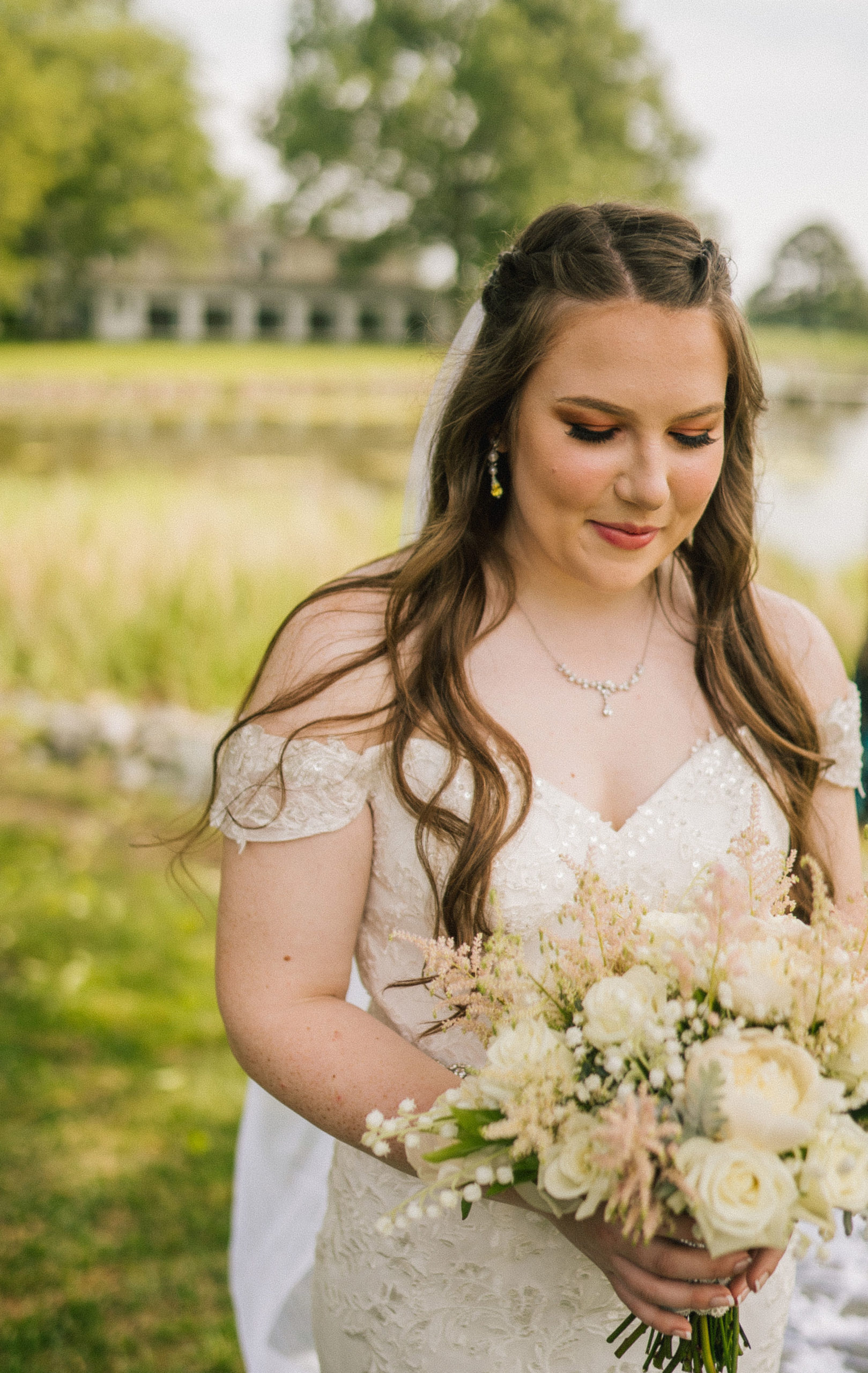 beautiful bride looking down at her white wedding bouquet as she smiles and stands next to the river at her wedding venue