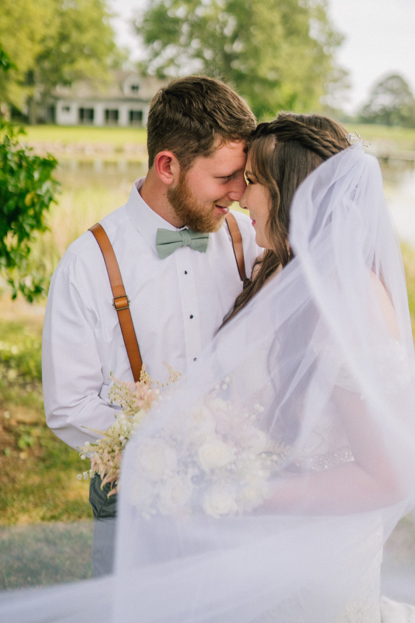 bride and groom hold their foreheads together as they embrace one another and the brides veil flows in front on them, bride wearing a lace mermaid gown and groom wearing suspenders and a mint bow tie