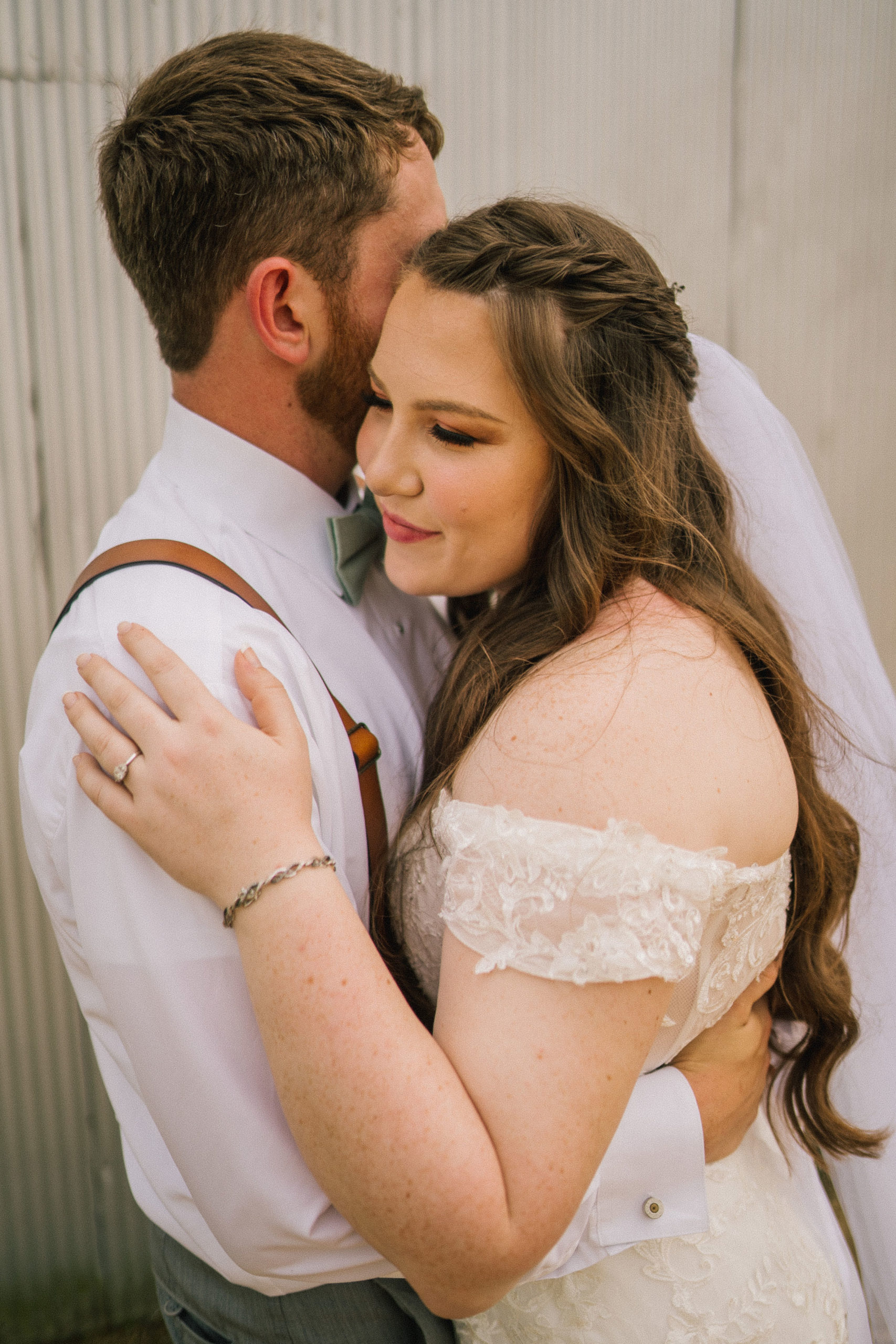 bridal portraits with groom holding his brides waist as she holds his shoulder in front of a grey barn in Maryland for their destination wedding day, while bride wears a stunning mermaid gown and long veil with lightly curled hair