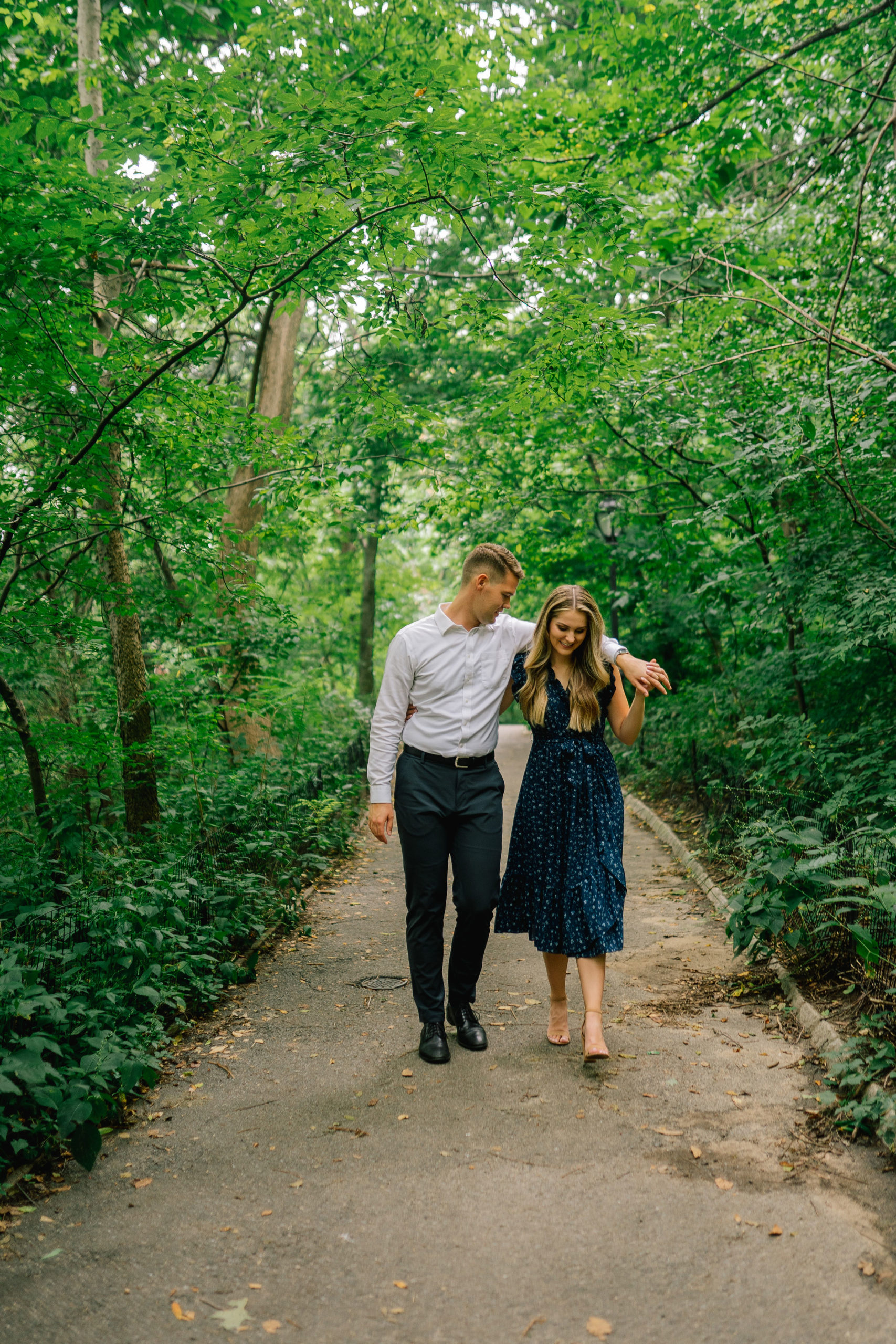woman in a dark blue dress walking with her fiance down a trail in Central Park for their engagement session with nest Brooklyn wedding photographer