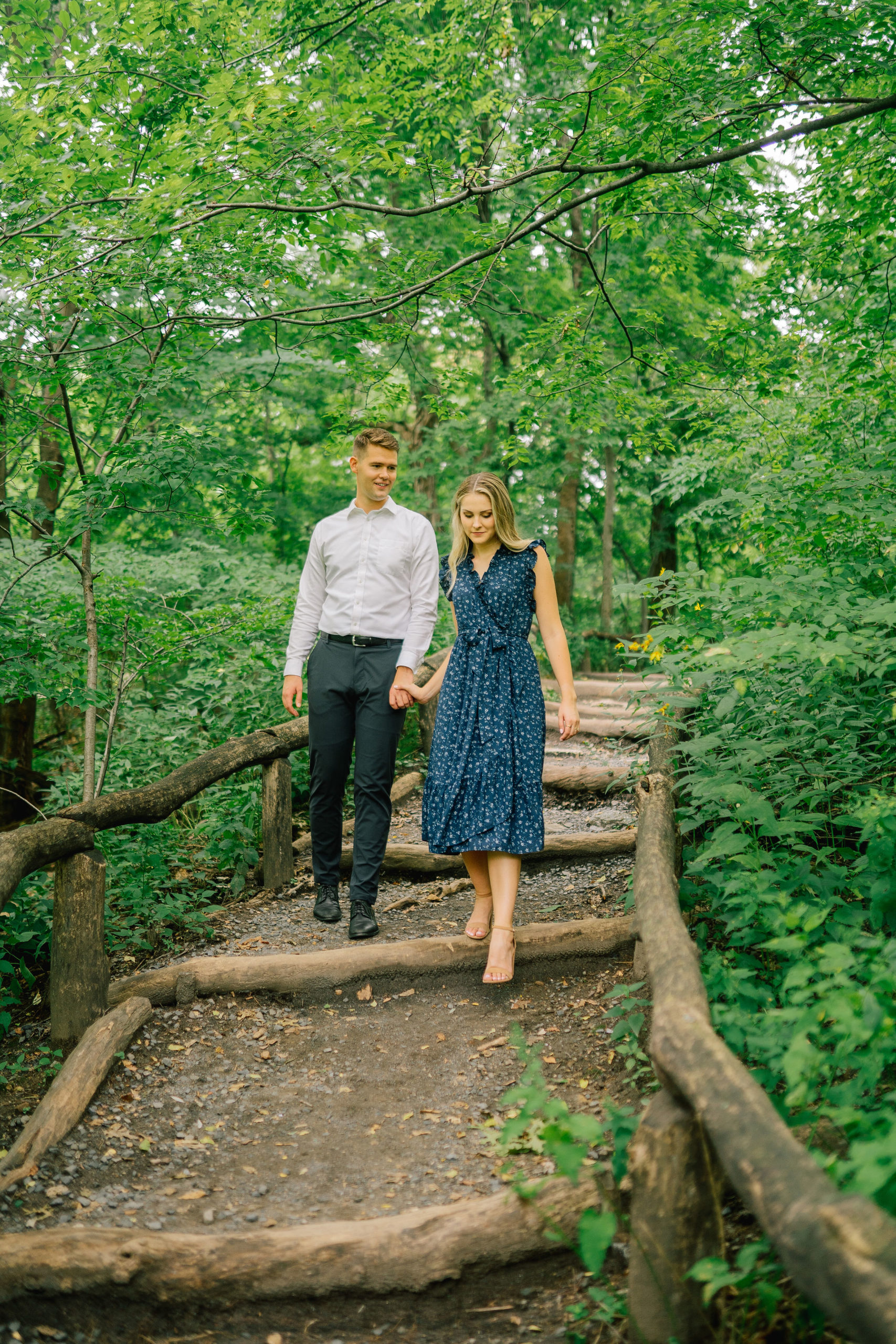 man and woman walking on a trail with small steps in Central PArk for their engagement session with bright green trees surrounding them as they holding hands and look down at the ground, moment captured by the best Manhattan wedding photographers
