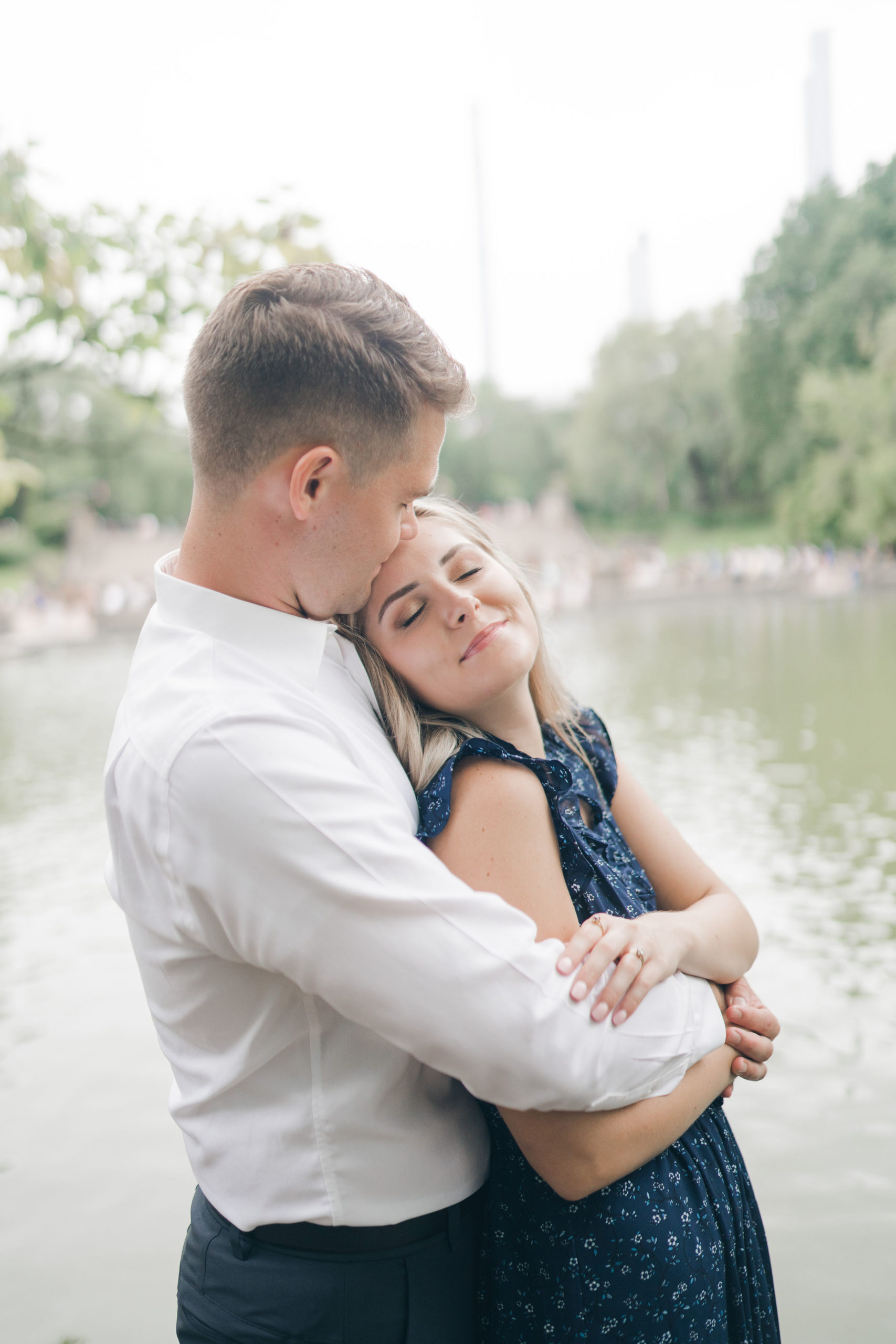 man standing behind his fiance in Central Park with his arms around her shoulders hugging her as she tilts her head back slightly and grins with a pond behind them captured by best Manhattan wedding photographers