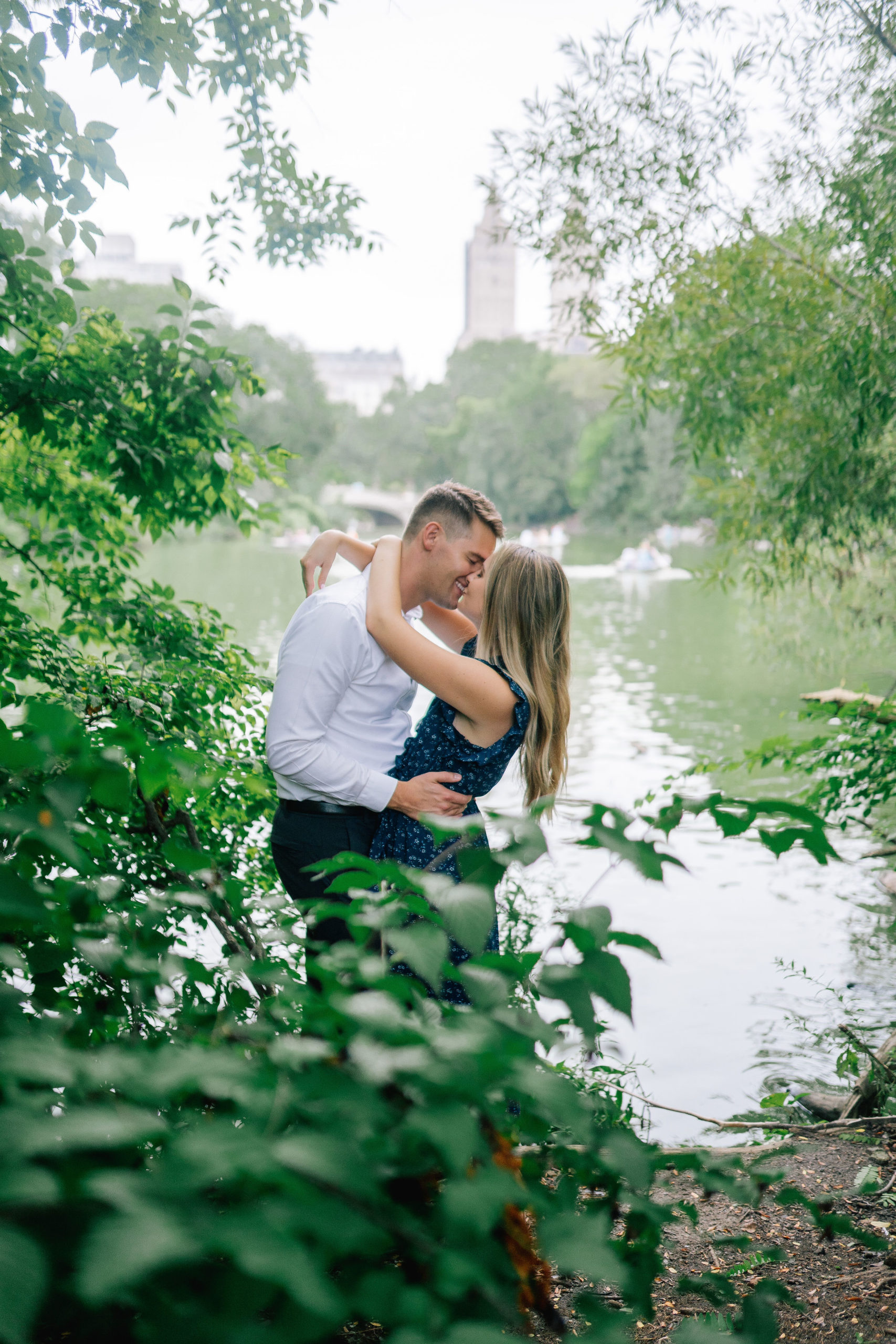 true loves kiss in the bushes in Central Park with a pond and the city in the back ground during an engagement photoshoot with best Manhattan wedding photographer