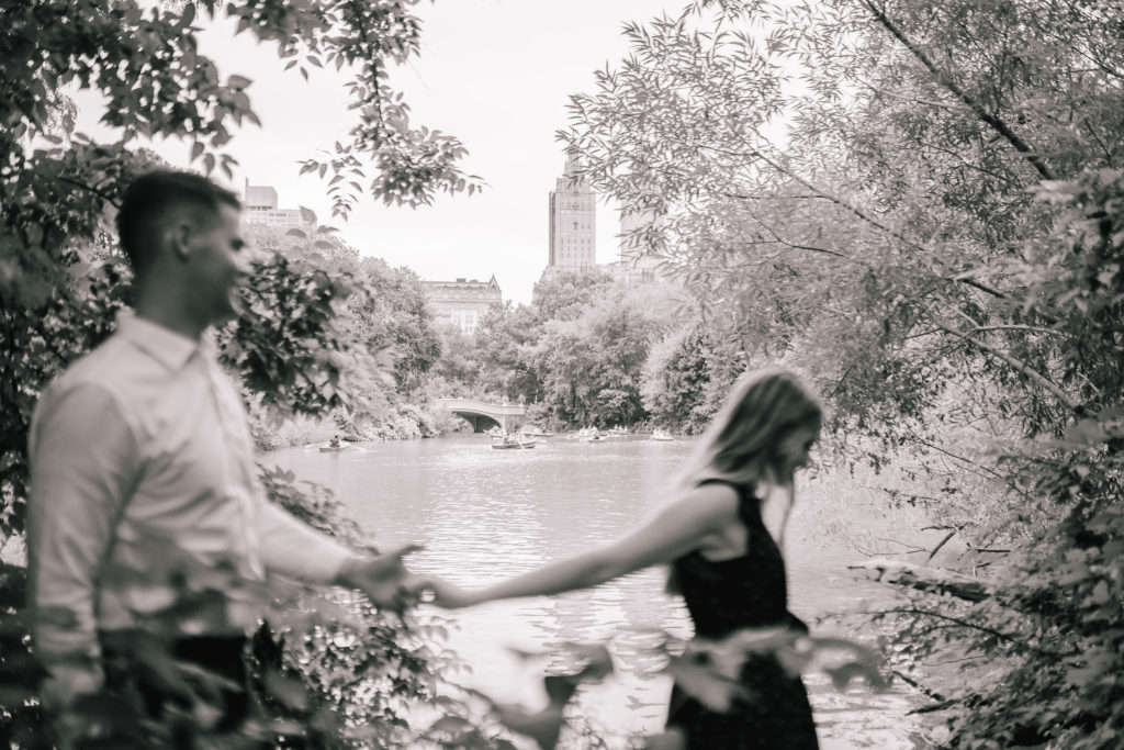 black and white image of woman and man holding hands as the woman walks in front of the man through some trees in Central Park with the skyline behind them