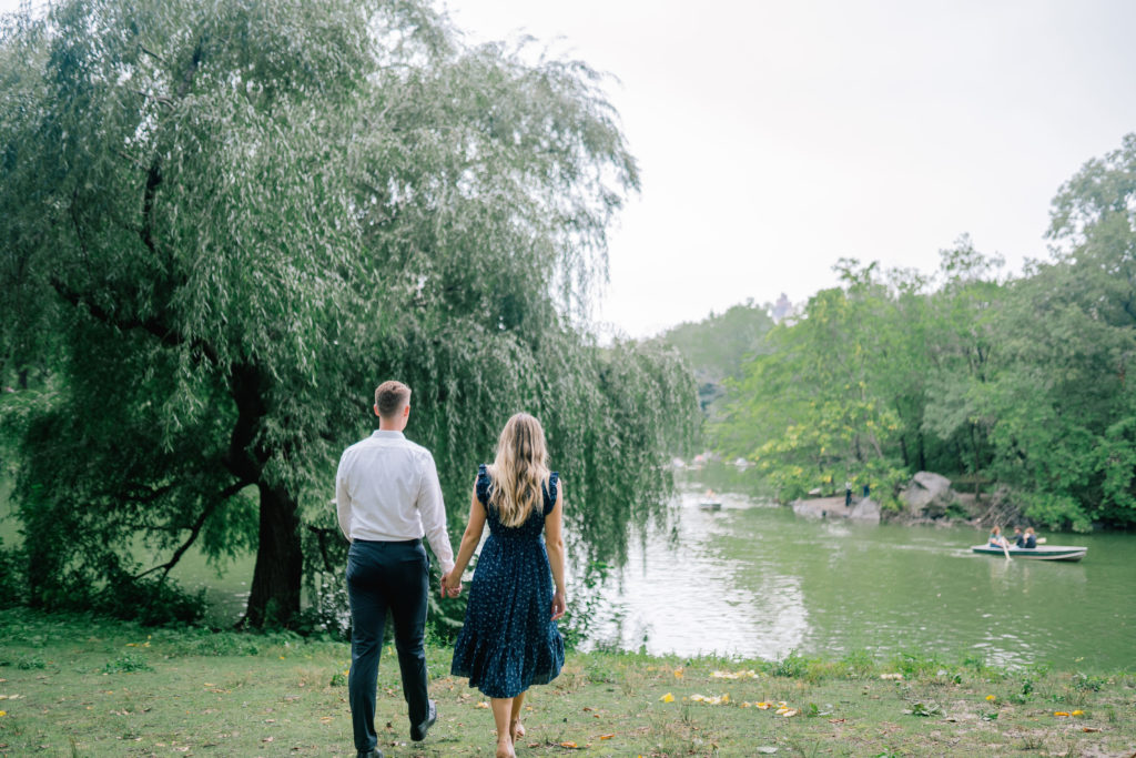 Manhattan engagement session in Central park with man and woman holding hands and walking towards a lake in Central Park next to a willow tree