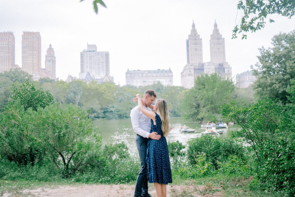 engaged couple dancing in their Central Park engagement photos next to a lake in New York City as they smile and the skyline is behind them