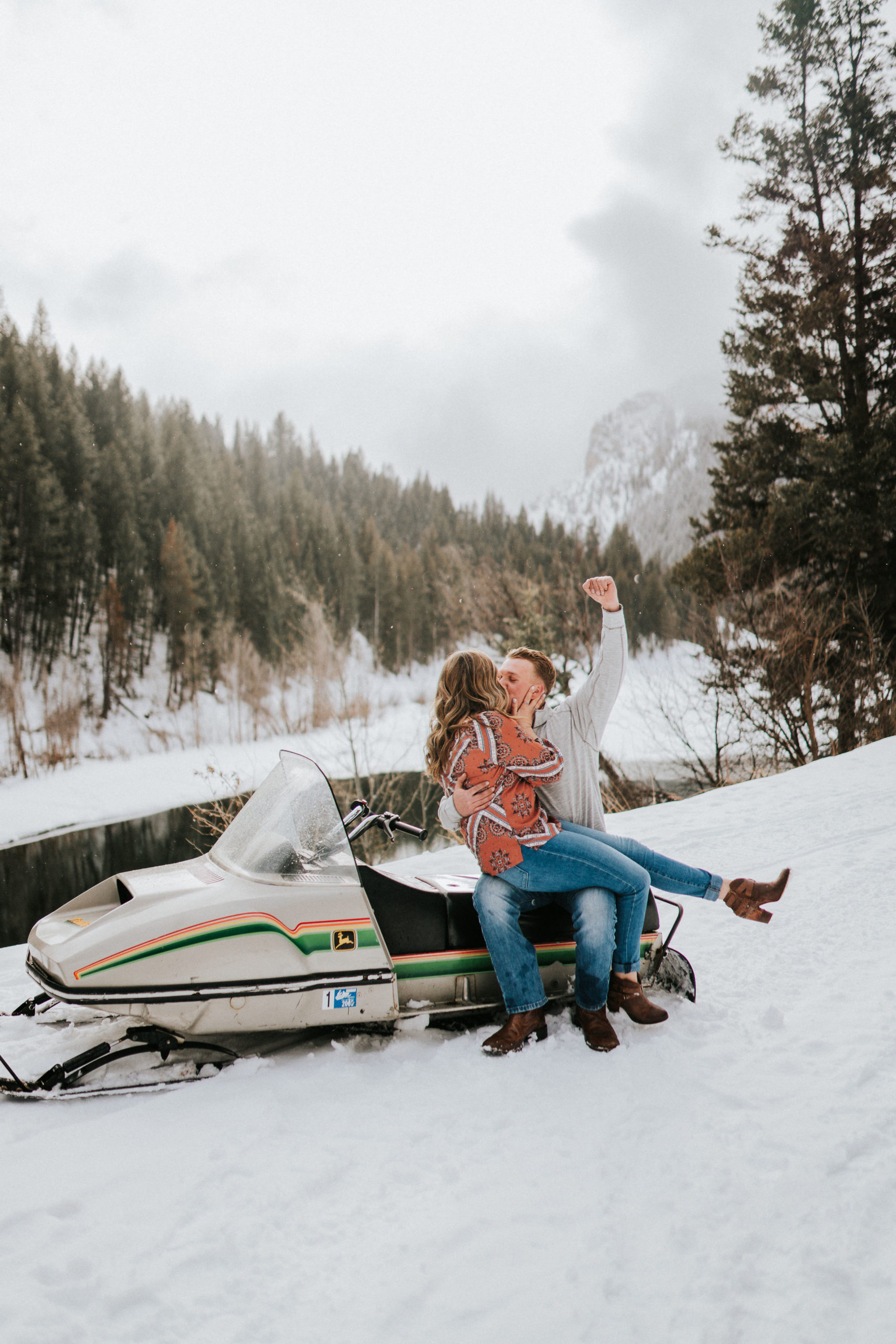 man and woman sitting on a snow mobile in the mountains surrounded by snow with the woman on the mans lap and the man raising a fist to the sky in victory as they kiss for their Smoky Mountain engagement photos