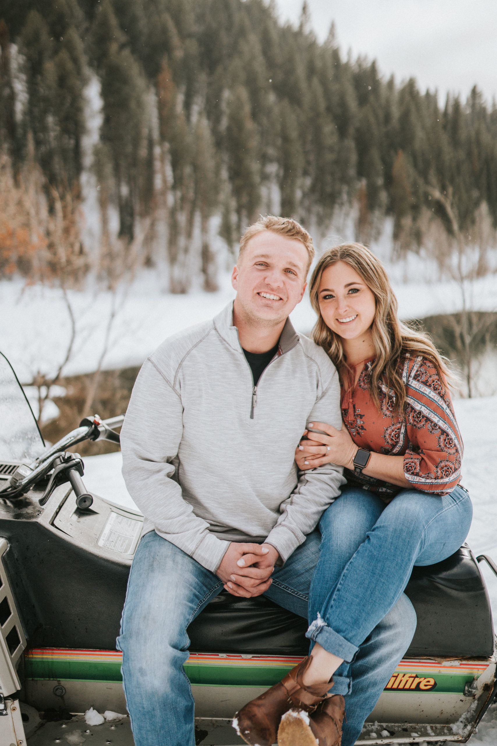 engaged couple sitting on a snow mobil ein the mountains and smiling for their winter engagement pictures