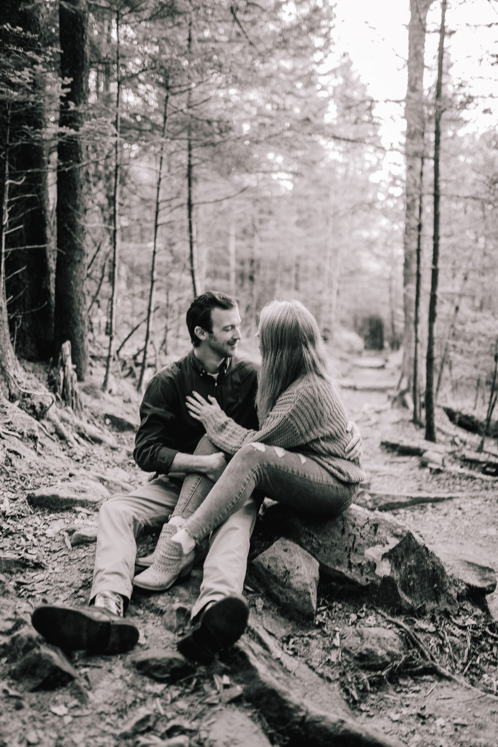 black and white engagement pictures with man and woman sitting on a rocky dirt trail in the Smokies with the woman holding the man by his chest as they both smile