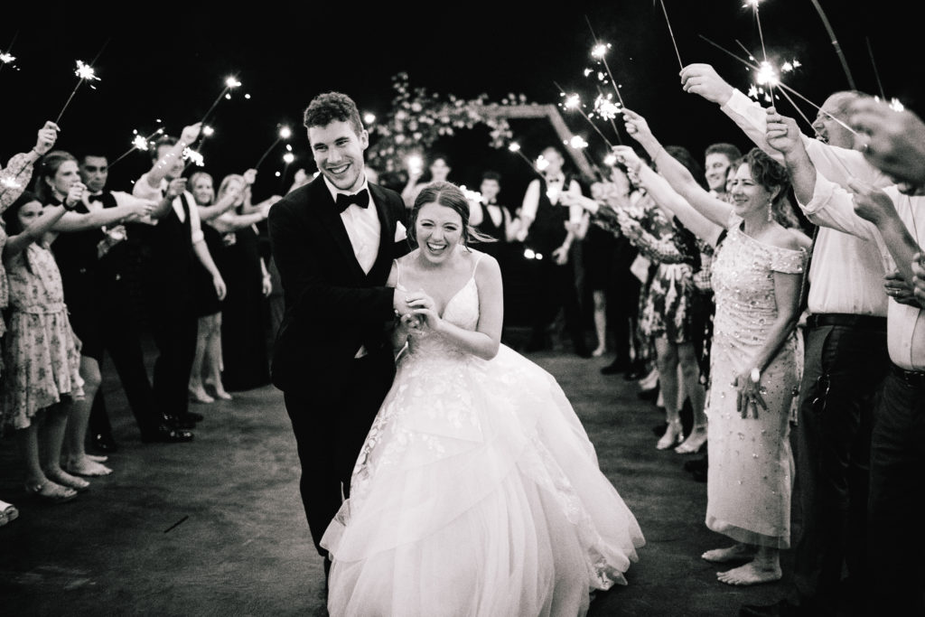 black and white image of bride and groom at the end of their wedding day during their send off with their guest and bridal party waving sparklers in the air