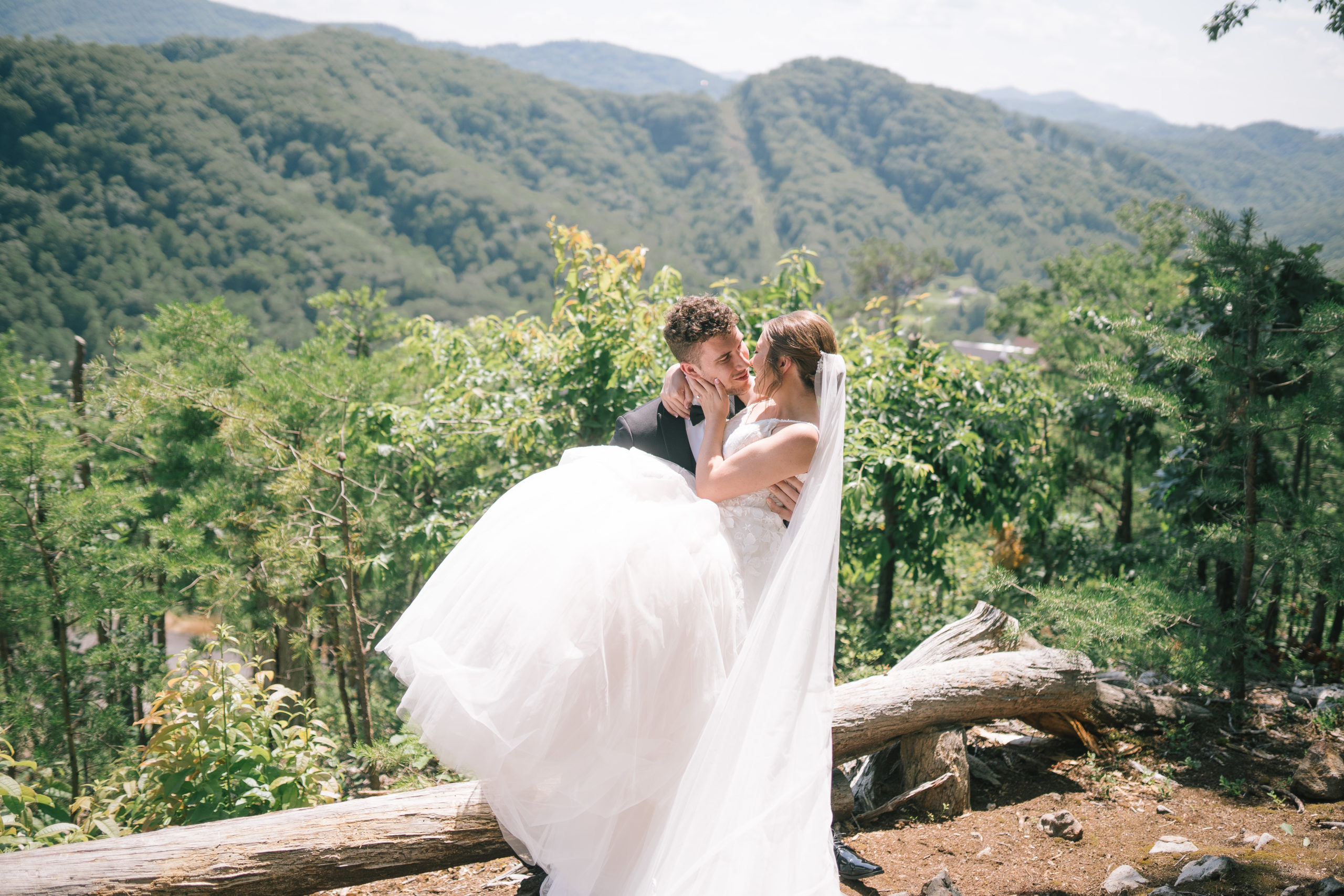 The Magnolia wedding venue in Tennessee with groom carrying his bride with the Smoky Mountains behind them for thier outdoor summer wedding