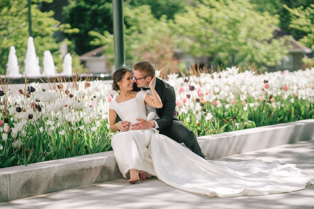 wedding photos on lds temple ground for Amalfi Coast themes wedding, mana and woman sitting on a bench in front of Spring flowers and holding each other 