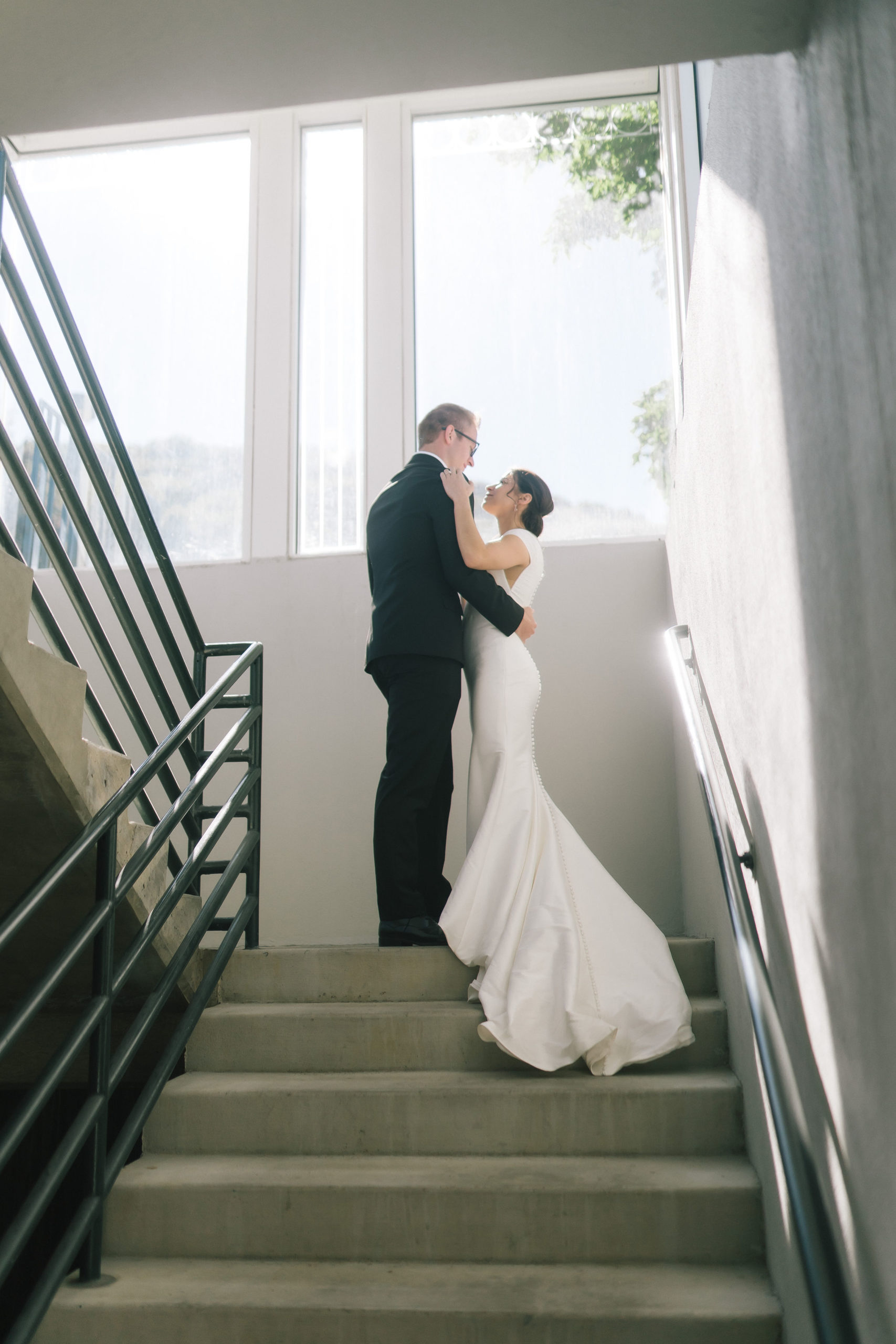 Amalfi Coast Themed Wedding Italian bride and groom standing in a stairwell on their wedding day and holding each other with a large bright window above them shining in the daylight