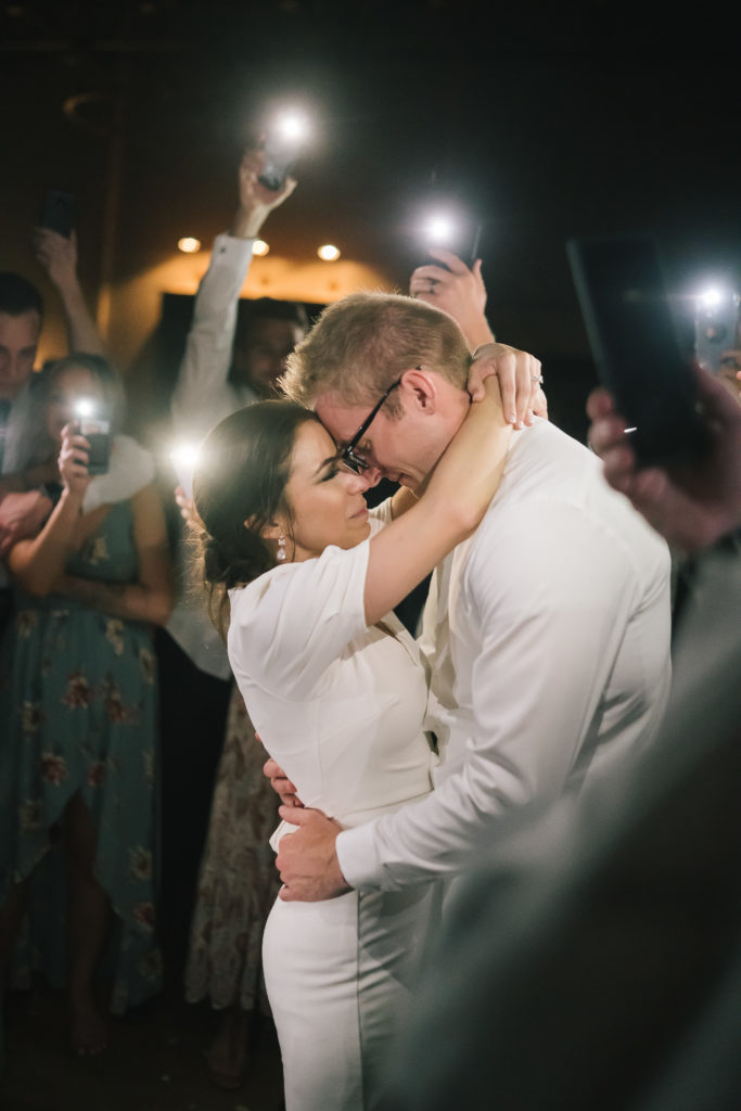 stunning wedding reception picture with everyone around the bride and groom holding their cell phone lights on as the bride and groom dances together